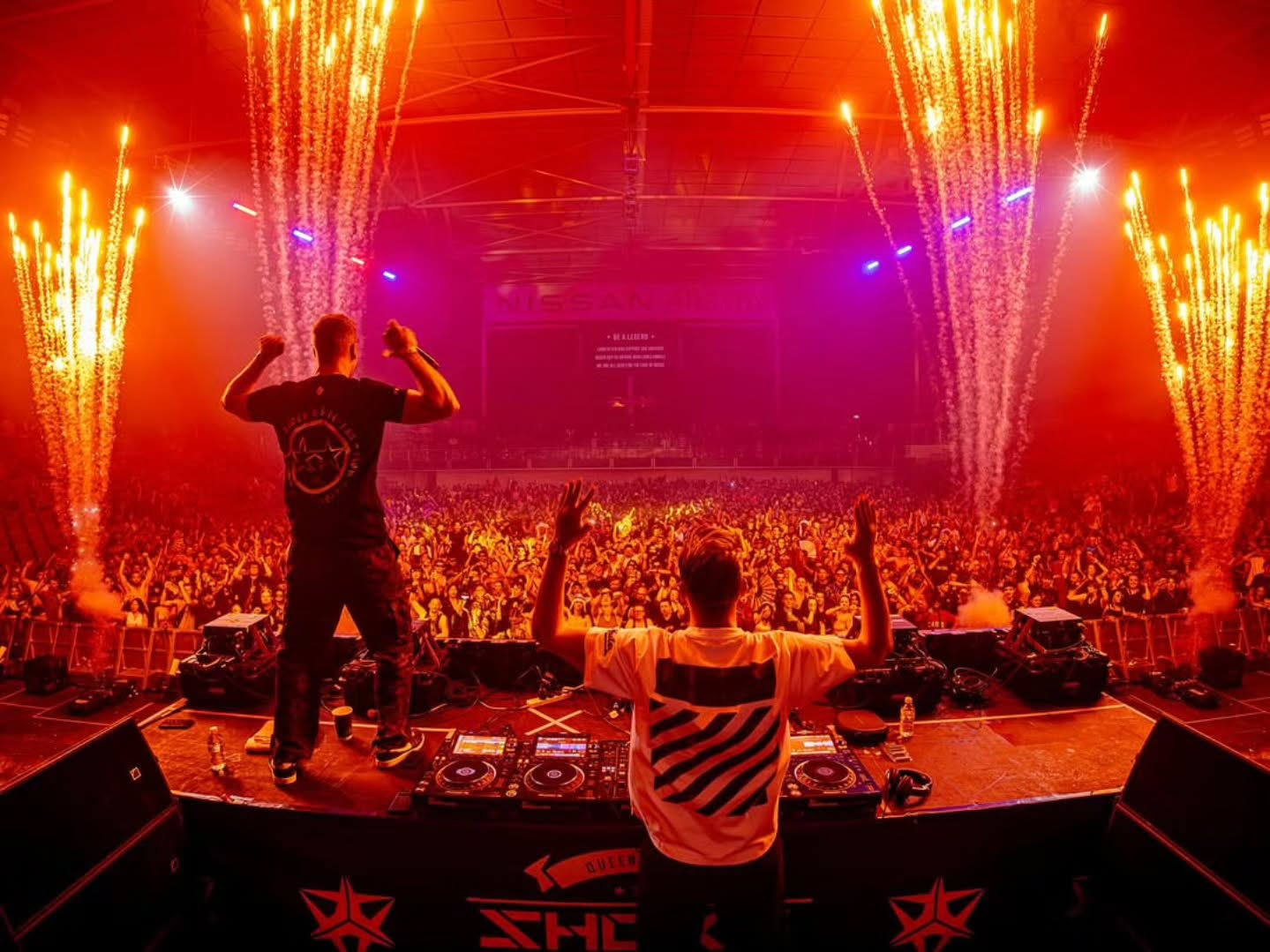 DJs performing on stage with close proximity fireworks either side under red lighting