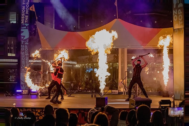 Fire performers with flame bursts on stage at a Winter City event, watched by a large crowd