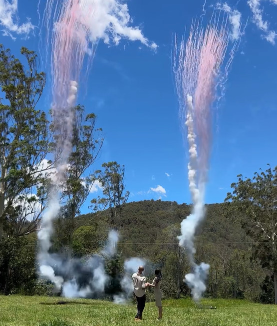 Couple standing together as pink coloured smoke daytime fireworks launch behind them at a gender reveal